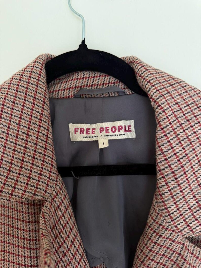 May include: Close-up of a coat with a red, white, and black plaid pattern. The coat has a gray lining and a label that reads "FREE PEOPLE". The coat is hanging on a black hanger.