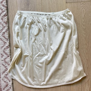 Op de afbeelding: Ivoor kleurige onderrok met een elastische tailleband en een delicate kanten rand langs de zoom. De rok is gemaakt van een gladde, zijdeachtige stof. Het kledingstuk is ontworpen om als onderkleding of als rok te worden gedragen.