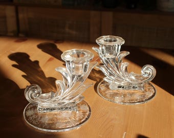 May include: Two clear glass candle holders with ornate designs. Each holder features a swirling, leaf-like pattern and a central cup for a candle.