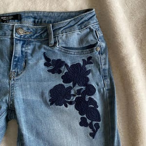 Puede incluir: Vaqueros de mezclilla azul claro con un diseño floral bordado en azul oscuro en la pierna derecha. Los vaqueros tienen una cintura azul oscuro con el texto "SIMPLY VERA" en blanco.