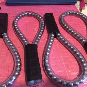 Op de afbeelding: Drie zwarte handvatten, zilverkleurige, flexibele lussen met een reeks metalen ballen erin. De lussen zijn gemaakt van een helder materiaal en zijn aan de handvatten aan elkaar gebonden.