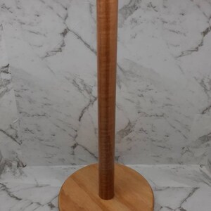 May include: A wooden paper towel holder with a round base and a cylindrical post. The holder is made of light brown wood.