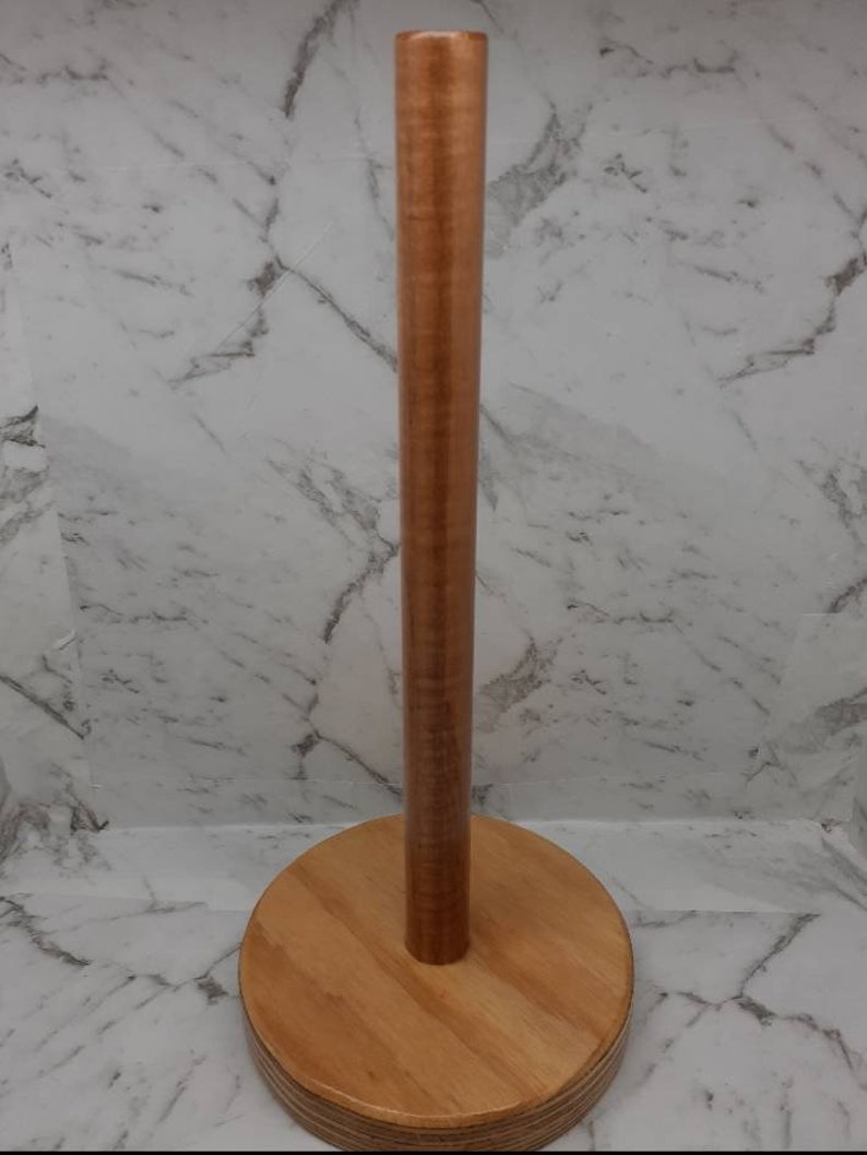 May include: A wooden paper towel holder with a round base and a cylindrical post. The holder is made of light brown wood.