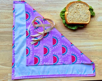 Puede incluir: Un envoltorio de comida de tela púrpura con estampado de sandía y forro blanco. El envoltorio está doblado en forma de triángulo y tiene una correa beige.