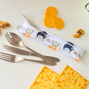 Op de afbeelding: Een witte ritszak met een luiaard- en regenboogontwerp, met een vork, mes, lepel en twee metalen rietjes. De zak ligt op een wit oppervlak met crackers en walnoten.