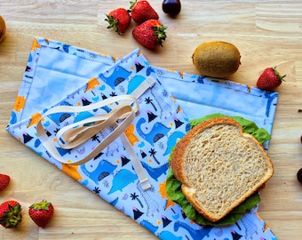 Puede incluir: Un envoltorio reutilizable para sándwich con un estampado de dinosaurios azules, que contiene un sándwich con lechuga. El envoltorio se asegura con una correa beige. Fresas y un kiwi sobre una superficie de madera.