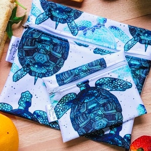 Könnte beinhalten: Drei wiederverwendbare Sandwichbeutel mit Reißverschluss, weißem Hintergrund und einem blauen und grünen Schildkrötenmuster. Die Beutel sind aus wasserdichtem Stoff gefertigt und eignen sich perfekt zum Verstauen von Snacks, Sandwiches und anderen Lebensmitteln.