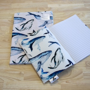 May include: A white notebook with a blue whale pattern cover. The notebook is open and has lined pages. The notebook is sitting on a wooden surface.