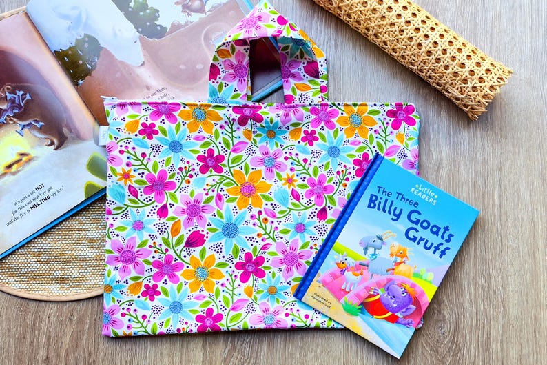 Puede incluir: Un bolso tote con estampado floral y asa a juego, junto a un libro infantil titulado "The Three Billy Goats Gruff". El bolso presenta un patr&oacute;n de flores rosas, azules y amarillas sobre un fondo blanco. Un libro abierto es visible.