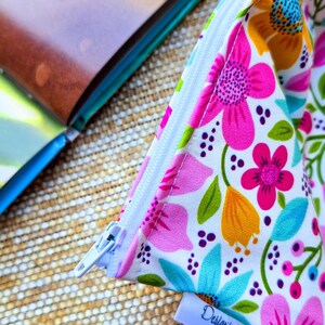 Puede incluir: Una bolsa con cremallera con estampado floral con fondo blanco, con flores rosas, naranjas y azules y hojas verdes. La bolsa est&aacute; abierta, revelando una cremallera blanca y una peque&ntilde;a etiqueta que dice "Design'r".