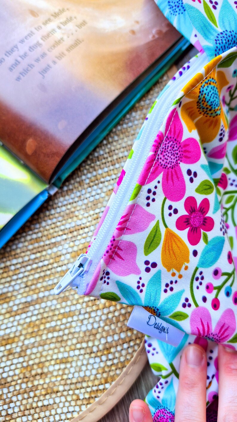 Puede incluir: Una bolsa con cremallera con estampado floral con fondo blanco, con flores rosas, naranjas y azules y hojas verdes. La bolsa est&aacute; abierta, revelando una cremallera blanca y una peque&ntilde;a etiqueta que dice "Design'r".
