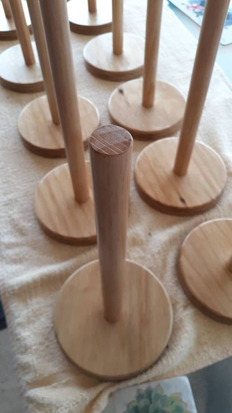 May include: Multiple wooden paper towel holders with a natural wood finish. The holders are round and have a cylindrical post in the center.