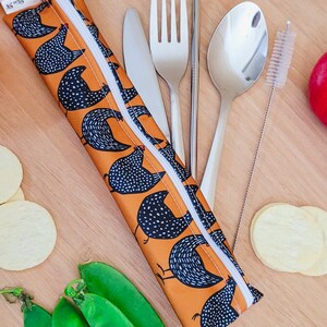 May include: An orange and white chicken-patterned utensil case with a white zipper, containing a knife, fork, spoon, chopsticks, and a straw cleaner. Fresh green pea pods and potato chips are arranged around the case on a wooden surface.
