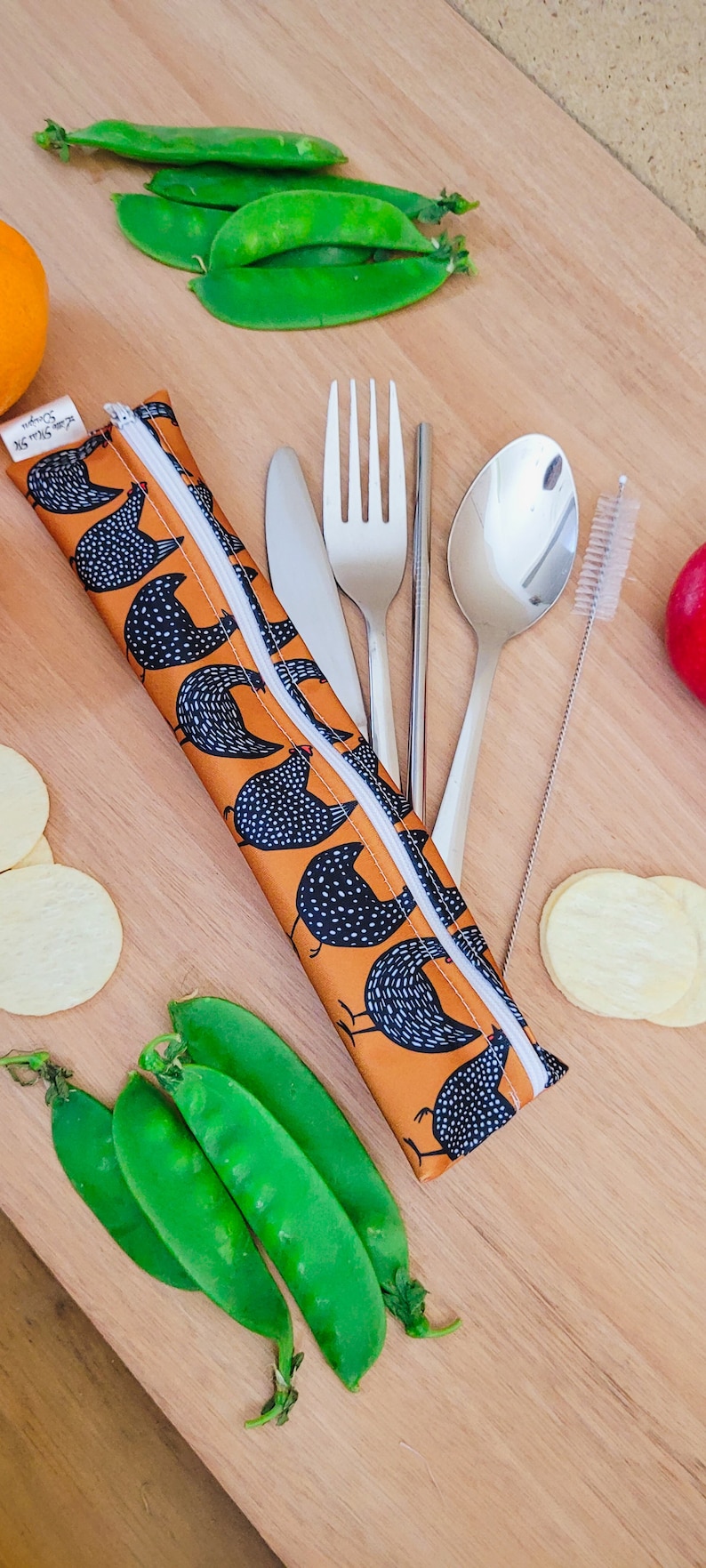May include: An orange and white chicken-patterned utensil case with a white zipper, containing a knife, fork, spoon, chopsticks, and a straw cleaner. Fresh green pea pods and potato chips are arranged around the case on a wooden surface.