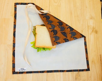May include: A brown and white fabric wrap with a black and white chicken pattern. The wrap is folded over a sandwich with lettuce and white bread. The wrap has a white label with the text "Bee's Wrap" on it.