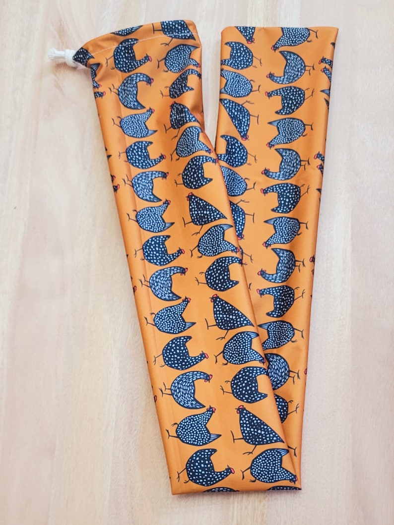 May include: Two long, orange fabric pouches with a repeating pattern of black and white speckled chickens. The pouches have a white drawstring.