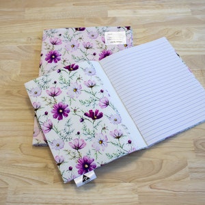 May include: Two notebooks with floral covers. The covers feature a white background with pink and purple flowers. The notebooks are open and show lined pages.
