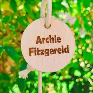 May include: A wooden keychain with the name "Archie Fitzgerald" engraved in a brown font. The keychain is round and attached to a silver clasp with a white rope. The background is a blurred green.