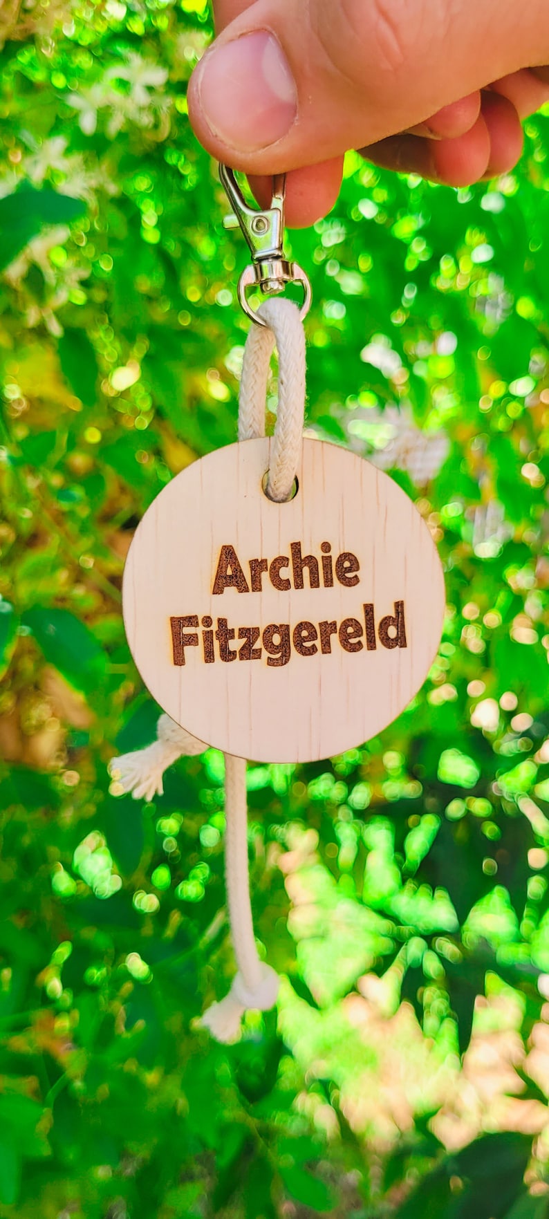 May include: A wooden keychain with the name "Archie Fitzgerald" engraved in a brown font. The keychain is round and attached to a silver clasp with a white rope. The background is a blurred green.