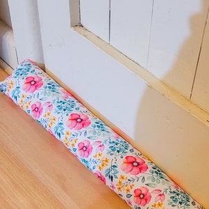 Könnte beinhalten: Ein weißer Türstopper mit einem floralen Muster mit rosa, gelben und blauen Blumen auf weißem Hintergrund. Der Türstopper liegt auf einem Holzboden.
