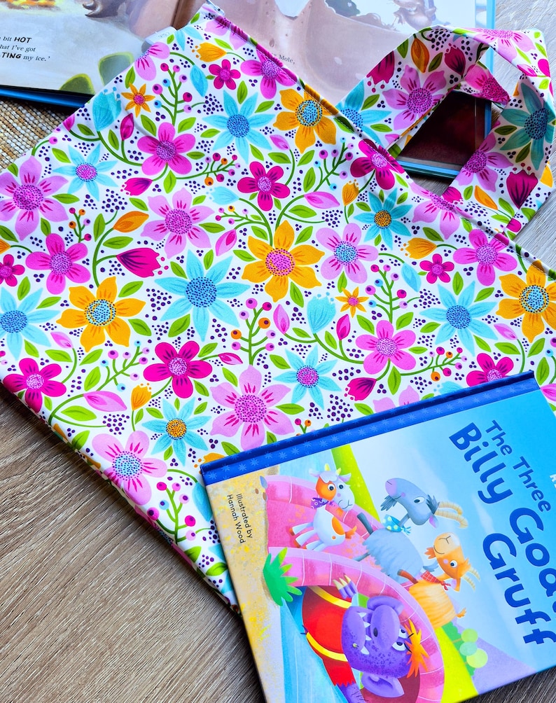 Puede incluir: Un bolso tote floral con un colorido estampado de flores rosas, azules y amarillas. Junto a &eacute;l, un libro infantil titulado "The Three Billy Goats Gruff". La portada del libro presenta ilustraciones de cabras y un trol.