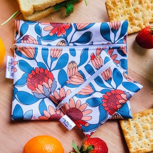 Può includere: Due sacchetti per la conservazione degli alimenti riutilizzabili con sfondo bianco e un motivo floreale con fiori rossi, arancioni e blu. I sacchetti hanno una chiusura a zip e il testo "Say No To Plastic" è stampato sui sacchetti.