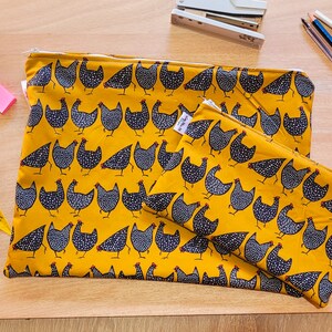 Può includere: Due pochette con cerniera in tessuto giallo con un motivo di pollo bianco e nero. La pochette più grande è sopra quella più piccola.