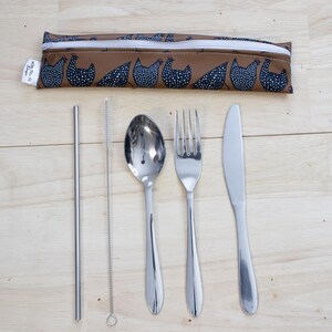 May include: A reusable cutlery set with a brown pouch featuring a chicken pattern. The set includes a stainless steel straw, straw cleaner, spoon, fork, and knife. The items are arranged on a light wood surface.
