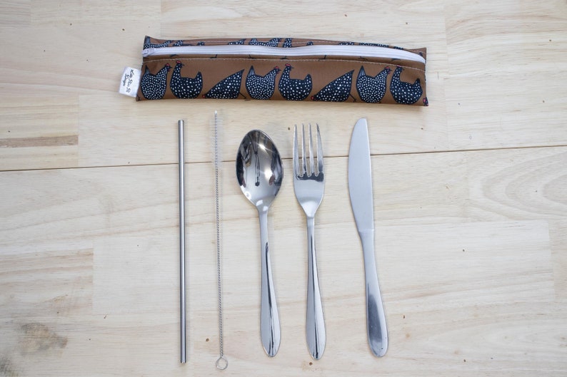 May include: A reusable cutlery set with a brown pouch featuring a chicken pattern. The set includes a stainless steel straw, straw cleaner, spoon, fork, and knife. The items are arranged on a light wood surface.