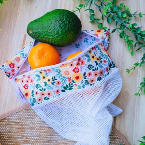 Könnte beinhalten: Eine weiße Netz-Obsttasche mit einer floralen Futter. Die Tasche ist gefüllt mit einer grünen Avocado und zwei orangefarbenen Mandarinen. Die Tasche liegt auf einer gewebten Matte.