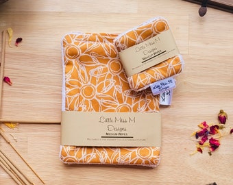 May include: Two reusable makeup remover wipes with a floral pattern in shades of yellow and white. The wipes are made by Little Miss M Designs and are labeled "Medium Wipes". The text "Reusable is the answer to a sustainable future" is printed on the bottom of the larger wipe.