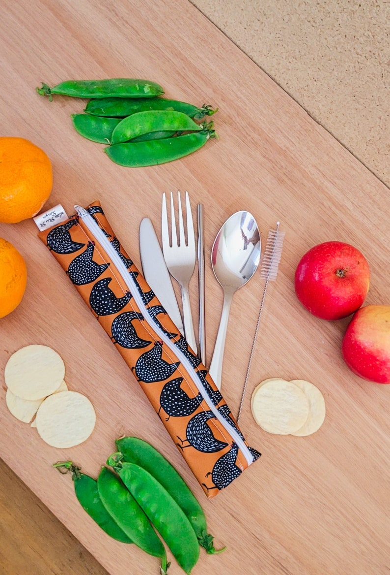 May include: A reusable cutlery set with a brown and black chicken print carrying case. The set includes a knife, fork, spoon, straw, and cleaning brush. Fresh green pea pods, oranges, apples, and chips are arranged around the set on a wooden surface.