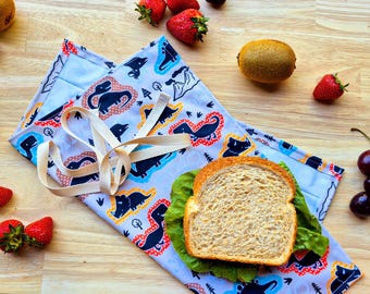 May include: A white fabric lunch bag with a dinosaur print and a brown strap. The bag is folded and a sandwich is inside. The bag is on a wooden surface with fruit and other food items.