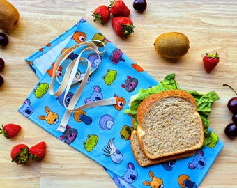 May include: A blue reusable sandwich wrap with a variety of animal illustrations, including koalas, kangaroos, and echidnas. The wrap is folded and has a white fabric lining. A sandwich is on top of the wrap, and there are cherries, kiwi, and strawberries scattered around.