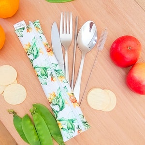 May include: A white zippered pouch with a sunflower pattern, containing a set of stainless steel cutlery: a fork, knife, spoon, and chopsticks. The pouch is lying on a wooden surface with a straw cleaning brush, two apples, and some crackers.