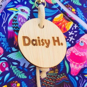 Puede incluir: Una etiqueta de madera con el nombre "Daisy H." grabado, sujeta a una bolsa con un colorido estampado de p&aacute;jaros. La bolsa tiene una cremallera y un cierre met&aacute;lico.