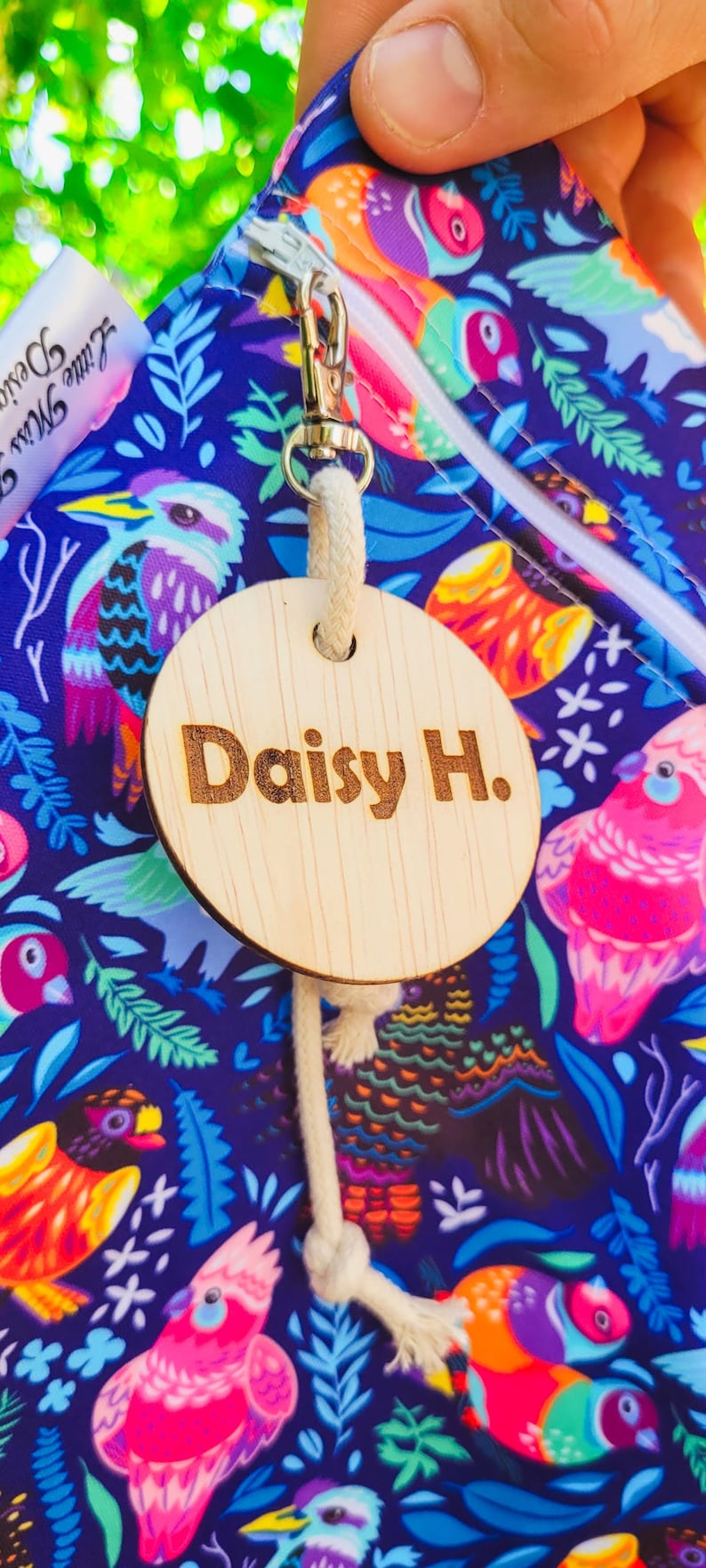 Puede incluir: Una etiqueta de madera con el nombre "Daisy H." grabado, sujeta a una bolsa con un colorido estampado de p&aacute;jaros. La bolsa tiene una cremallera y un cierre met&aacute;lico.
