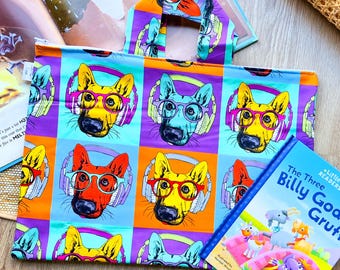 May include: A colorful tote bag with a repeating pattern of cartoon dogs wearing headphones and glasses. The bag has a purple, orange, and blue color scheme. The bag is lying on a wooden surface with a children's book titled "The Three Billy Goats Gruff" in front of it.