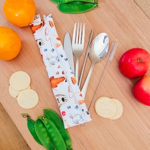 May include: A white zippered pouch with a cartoon dog print, containing a fork, knife, spoon, and a straw cleaning brush. The pouch is on a wooden surface with green pea pods, oranges, and round crackers.
