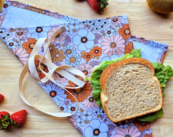 Puede incluir: Una bolsa de almuerzo de tela con estampado floral, una correa blanca y un sándwich en su interior. La bolsa está doblada por la mitad y tiene un fondo azul claro con flores rosas, naranjas y blancas.