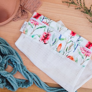 Könnte beinhalten: Eine weiße Netz-Obst- und Gemüsetasche mit einer floral bedruckten Reißverschlusstasche. Die Tasche hat einen weißen Hintergrund mit rosa, orangefarbenen und grünen Blumen. Der Text "The Flower Garden" ist auf der Tasche aufgedruckt.