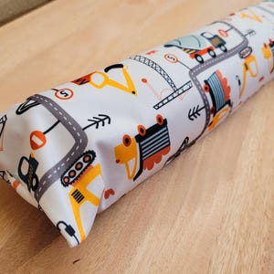 Könnte beinhalten: Ein weißer Türluftstopper aus Stoff mit einem farbenfrohen Muster von Baufahrzeugen. Das Design umfasst Straßen, Pfeile und verschiedene Baufahrzeuge in Orange, Gelb, Grau und Blau. Der Stopper liegt auf einer Holzoberfläche.