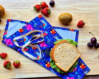 Puede incluir: Un envoltorio de comida reutilizable azul y blanco con un patrón de pájaros coloridos. El envoltorio está doblado alrededor de un sándwich con lechuga y una rebanada de pan blanco. El envoltorio tiene una correa blanca para llevar.