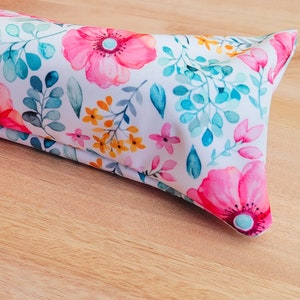 Könnte beinhalten: Ein weißes Stoffkissen mit einem Blumenmuster mit rosa, gelben und blauen Blumen. Das Kissen ist in der Hälfte gefaltet und liegt auf einer Holzoberfläche.