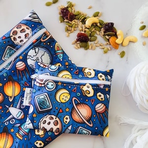 Può includere: Due sacchetti per snack riutilizzabili con sfondo blu e stampa colorata a tema spaziale. I sacchetti hanno una chiusura a zip e il testo "Little Plus Designs" è stampato sul sacchetto più grande.