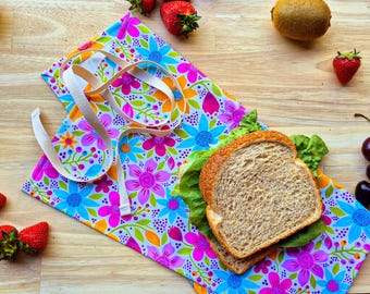 Puede incluir: Una bolsa de almuerzo de tela floral colorida con una correa marrón y un sándwich dentro. La bolsa está sobre una superficie de madera con fruta y otros alimentos.
