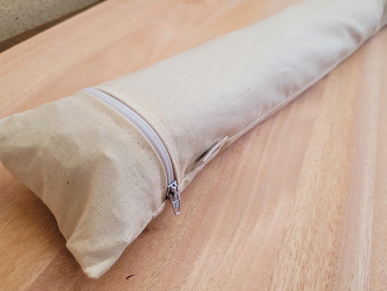 May include: A beige canvas zippered pouch or bag. The zipper is white and the pouch is lying on a light brown wooden surface.