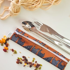 May include: A reusable utensil set with a brown chicken-print carrying case, stainless steel spoon, fork, knife, and straw. Trail mix is scattered around the set. A macrame wall hanging is in the background.