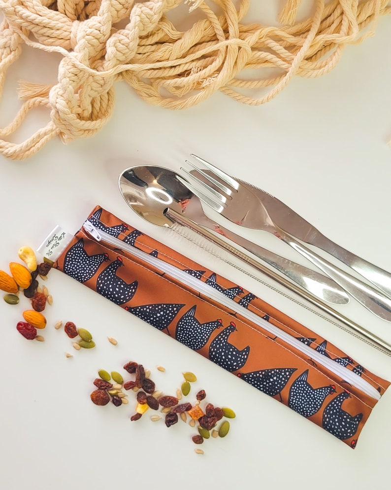 May include: A reusable utensil set with a brown chicken-print carrying case, stainless steel spoon, fork, knife, and straw. Trail mix is scattered around the set. A macrame wall hanging is in the background.