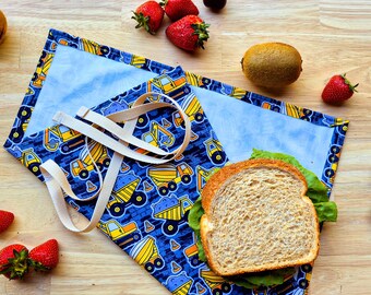 May include: A blue and yellow reusable lunch bag with a construction vehicle print. The bag has a white strap and is folded over a sandwich.